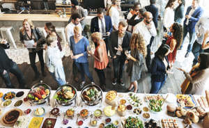 Feeding employees. A diverse group of people standing around talking and eating. A long table with a white linen is at the bottom of the image. There are a lot of diverse food dishes there as a buffet.