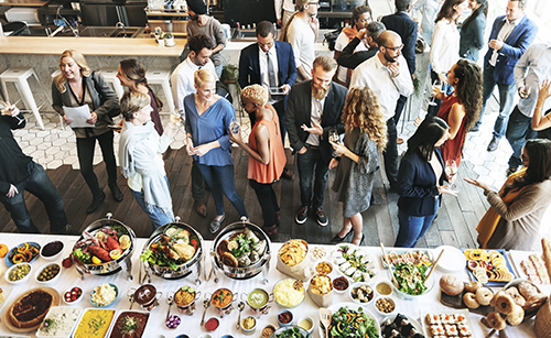 Feeding employees. A diverse group of people standing around talking and eating. A long table with a white linen is at the bottom of the image. There are a lot of diverse food dishes there as a buffet.