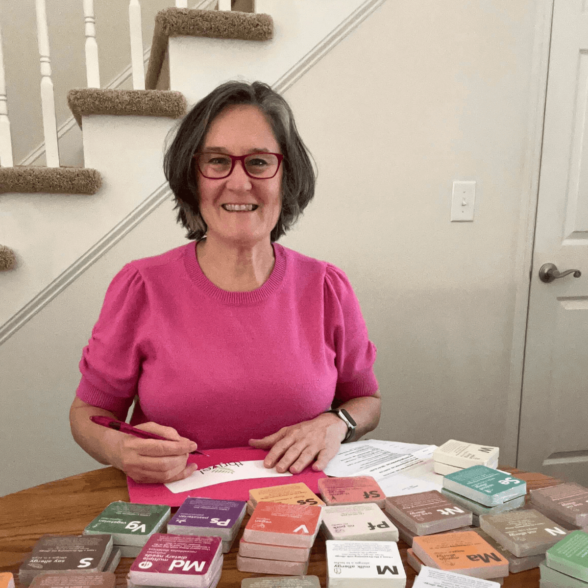 Smiling woman sitting at a table wearing a shortsleeve pink sweater. On the table in front of her are piles of dietary need meal cards in a variety of colors and options.