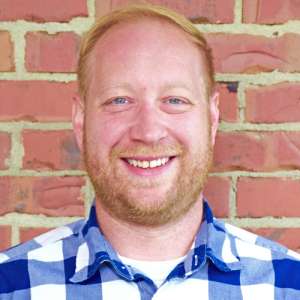 Ben Chapman - smiling white man with red hair and beard in blue & white checkered shirt standing in front of a brick wall