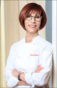 Female chef in her white coat leaning on a wall. She is wearing glasses and smiling. Fran Costigan vegan desserts