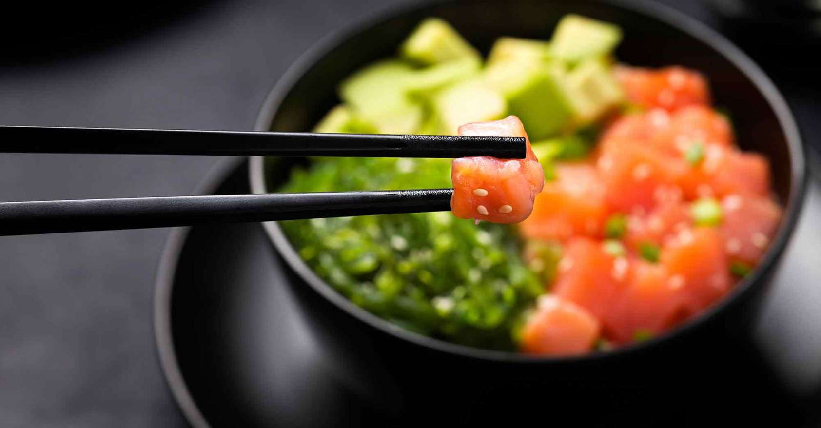 Poke with diced red fish avocado and green seaweeds salad with piece of salmon in chopsticks in foreground 2020 trends