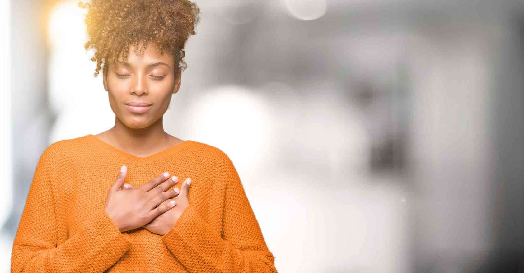 black woman in orange sweater with her hands on her heart self care