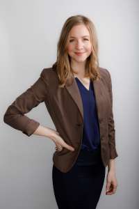 Woman with long blonde hair standing tall wearing a blue shirt and brown jacket with her right hand on her hip she promotes food integrity
