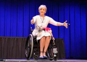 woman in a wheelchair on a stage holding her arms out forward. Her service dog is in her lap. She talks about how to be inclusive for Those with Disabilities