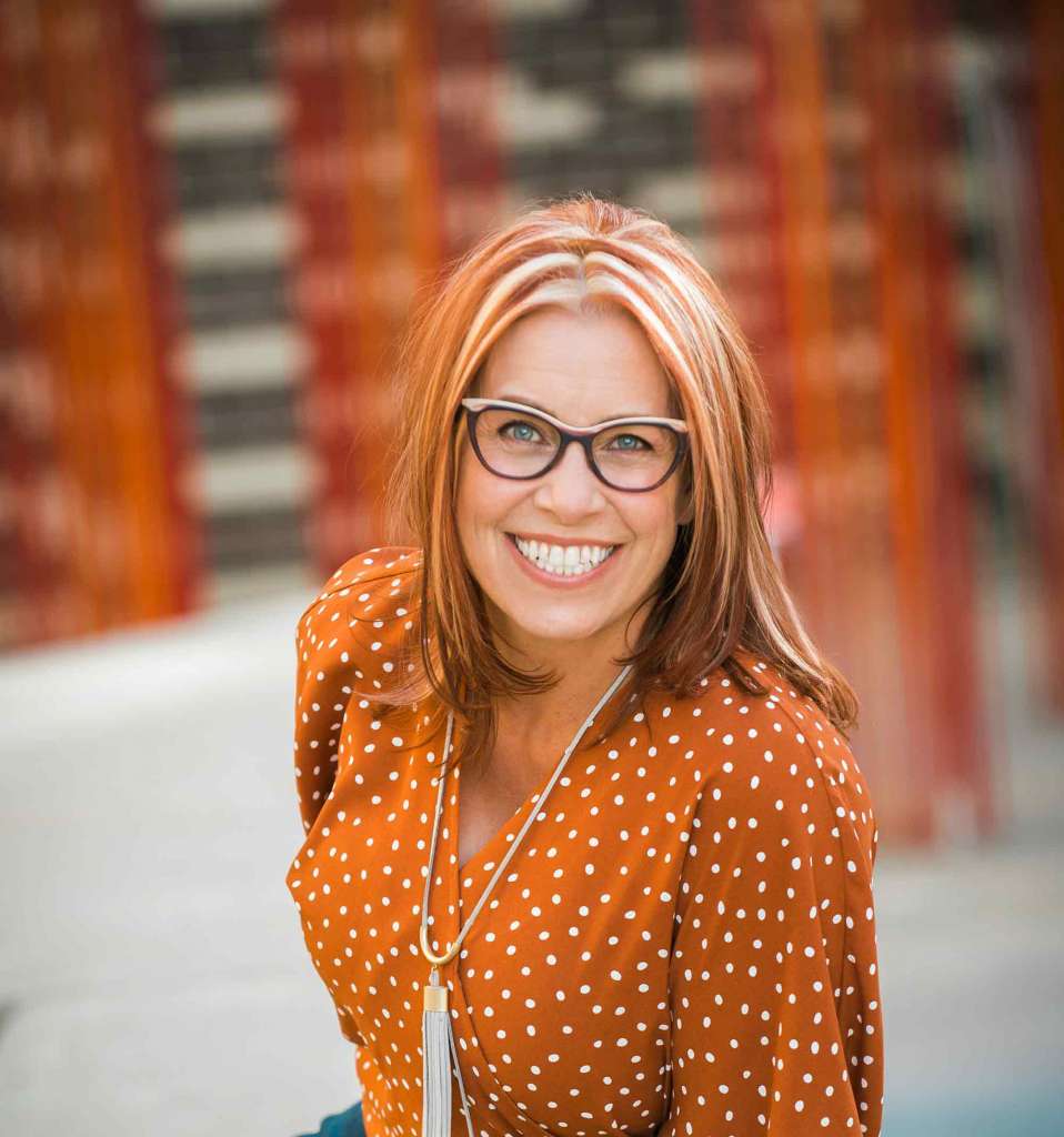 Smiling red headed woman leaning towards the camera in an orange blouse with white dots. She works to Foster Inclusive Workplace Culture
