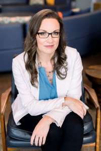 Woman in a blue blouse and white jacket leaning forward with arms crossed on her legs. She works to Elevating Food Allergy Safety