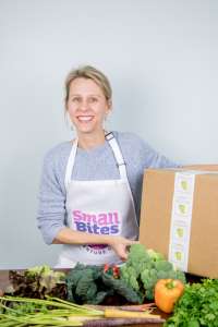 smiling woman behind a table full of vegetables and holding a box; CEO of Small Bites Adventure Club