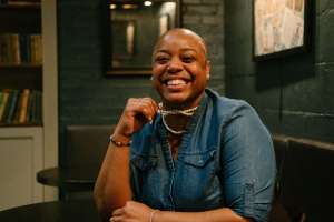 smiling woman in a denim shirt leaning on a table holding her necklace in her hand Barriere