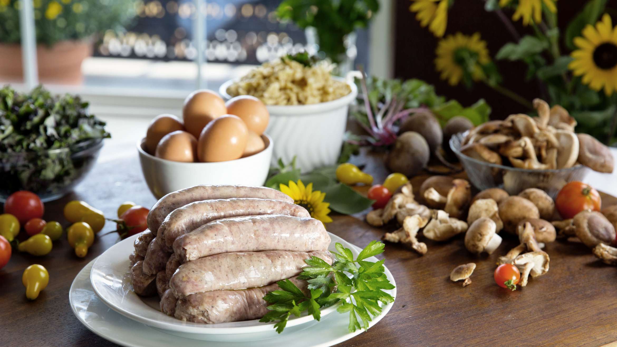 Plate with a stack of sausage on a wooden table with a bowl of eggs behind them and vegetables scattered around them