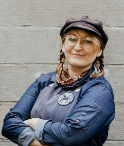 Smiling woman in a chef's blue coat and hat. Her hair is in braids and arms are crossed. She honors her tribal heritage