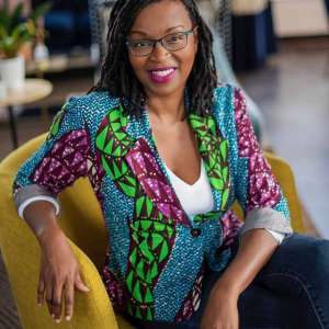 Smiling woman sitting in a yellow chair with her arm on the back of the chair wearing a colorful jacket which looks like paradise