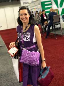 Smiling woman with long black hair in a purple dress with the words Sustainable Development Goals on the front. She is carrying a purple bag as well. Both were made from recycled signage showcasing sustainability efforts by a conference. She works for the Events Industry Council
