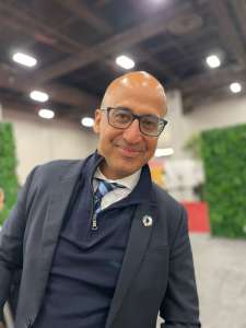 A baldheaded smiling man with glasses who is leaning to the right in front of green sustainability walls. He chairs the She works for the Events Industry Council Sustainability Committee