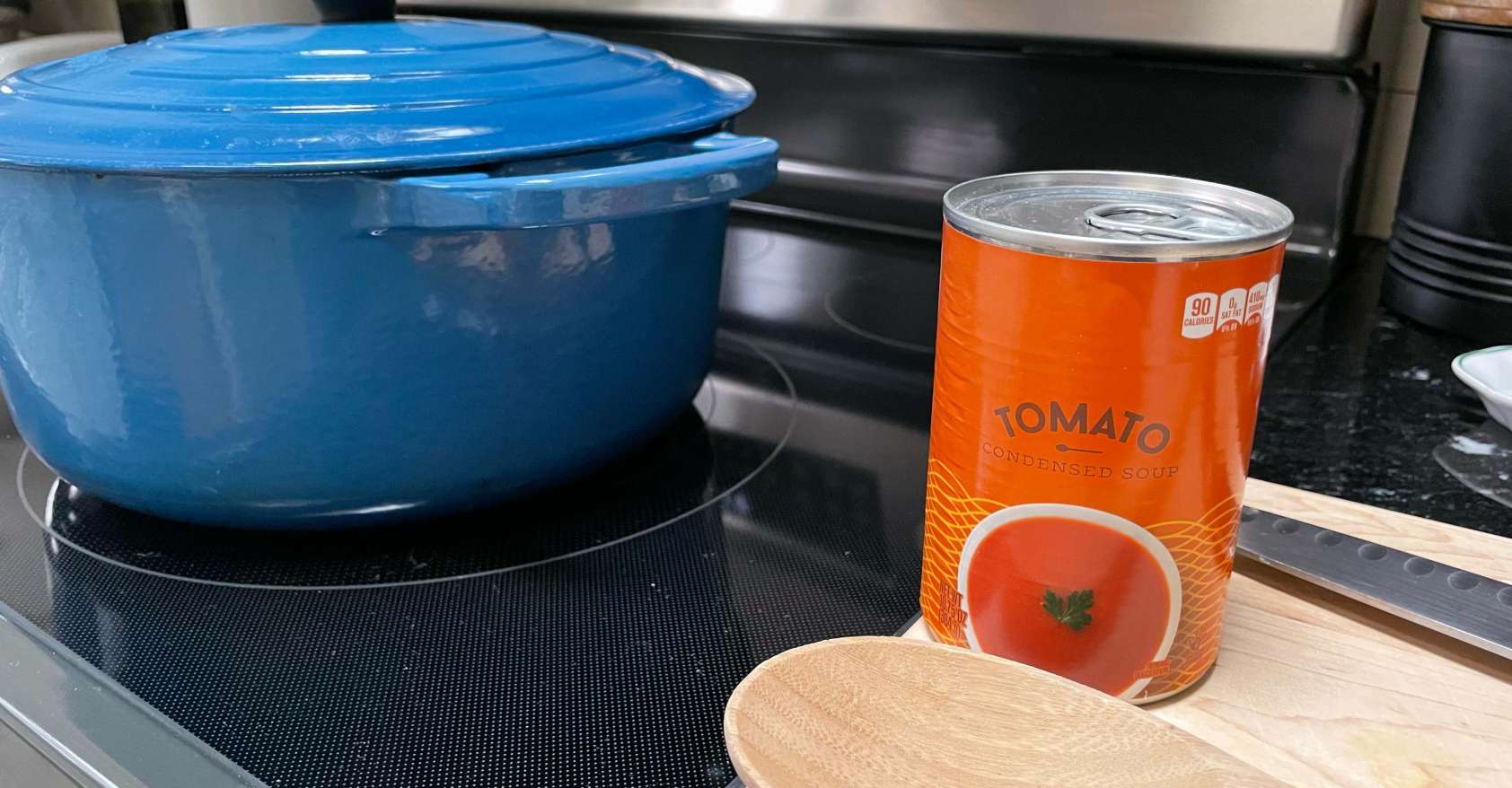 a can of tomato soup to be used for stew sitting on a cutting board with a wooden spoon next to stove with a large blue pot