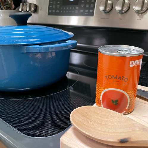 a can of tomato soup to be used for stew sitting on a cutting board with a wooden spoon next to stove with a large blue pot