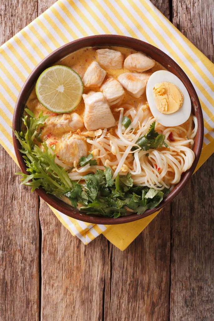 Malaysian laksa soup with chicken, egg, noodles and herbs close up in a bowl. Vertical top view 2022
