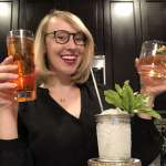 Smiling woman wearing glasses and holding a cocktail in each hand