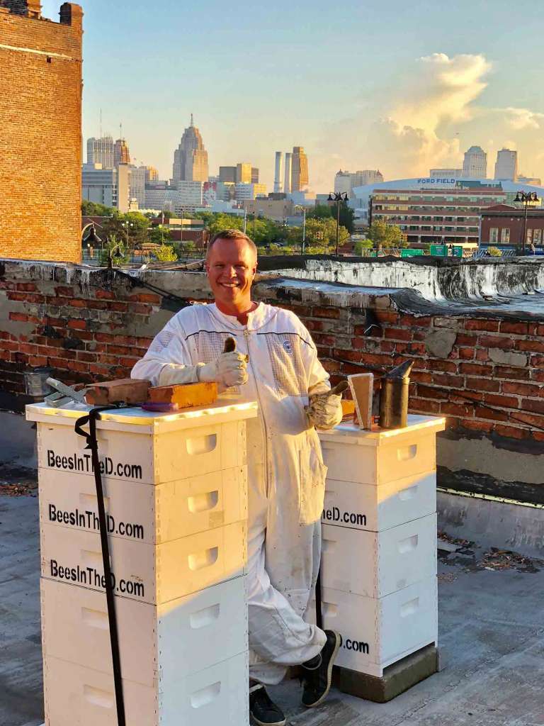 Beekeeper Brian Peterson leaning on bee hive on top of building. The city of Detroit behind him