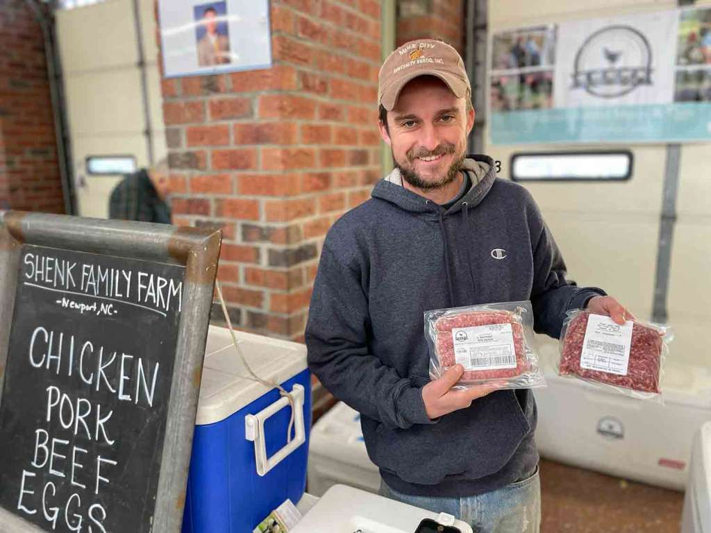 Smiling man holding two packages of frozen pork. A sign to the left of him says Shenk Family Farm Newport, NC with the words Chicken, Pork, Beef and Eggs in all capitals.