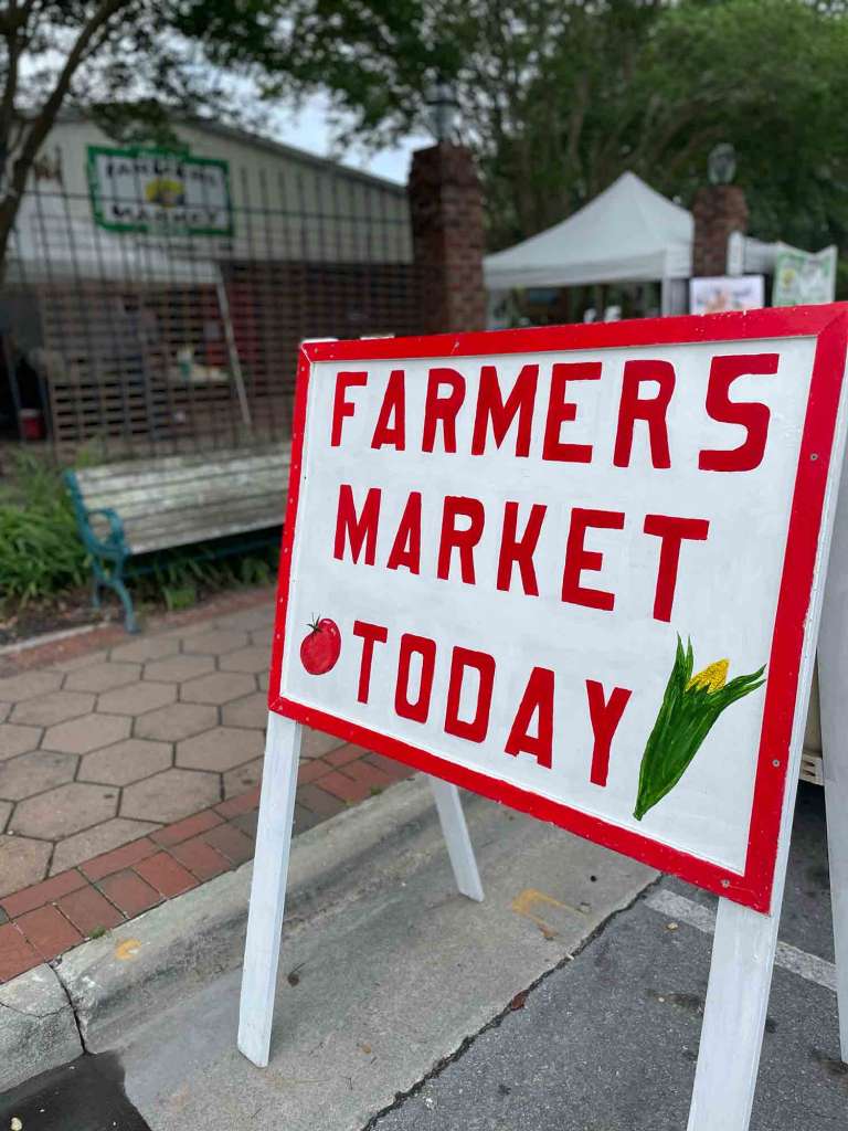 A wooden A-frame sign with four legs painted white. There is a red frame around the top rectangle sign. In the center are the words Farmers Market Today. A painted tomato is on the left of the word today and an ear of corn to the right. The New Bern Farmers Market has farmers who sell what's in season on their farms.