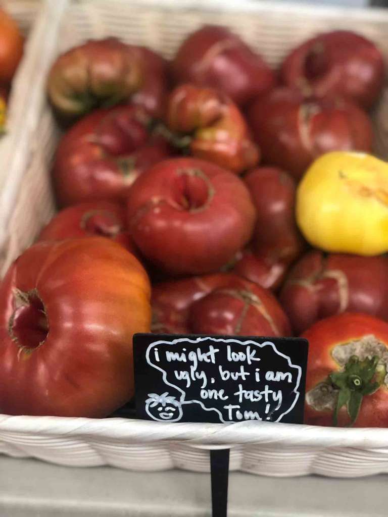 A basket of heirloom tomatoes. A small black sign at the front of the basket says "I might look ugly, but I am one tasty Tom!". 