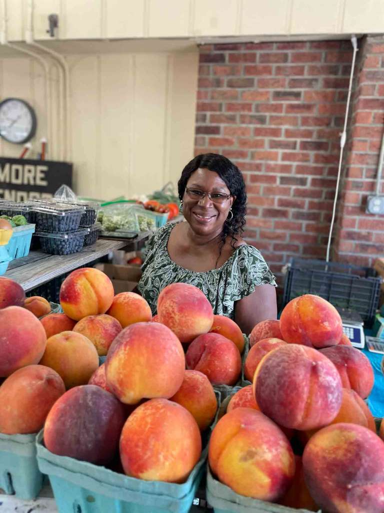 Smiling woman standing behind a table full of peaches in green cartons. Behind her is a brick wall. 