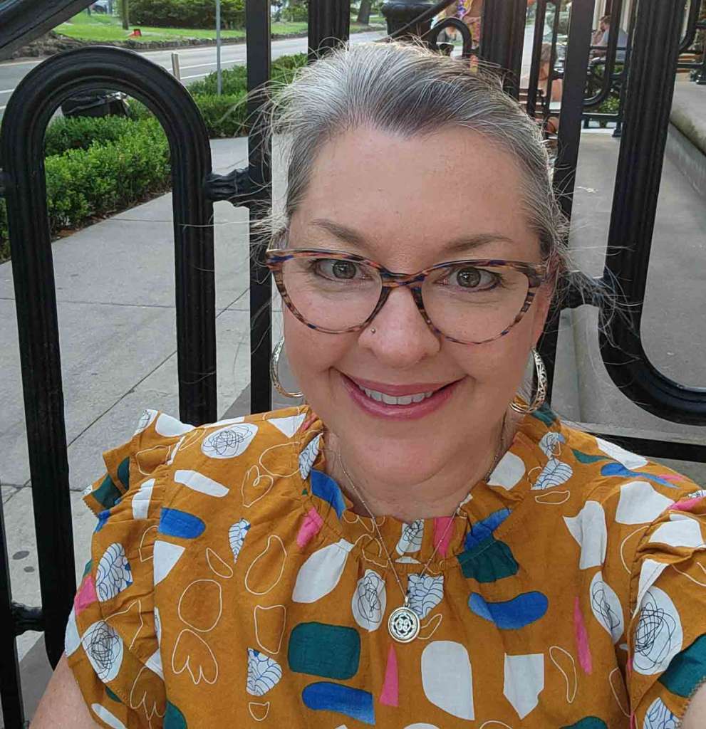 Smiling woman wearing glasses and orange shirt with blue and white geometric objects. She is sitting outside. Aurora Dawn Benton, Sustainability champion. Safe, Sustainable & Inclusive