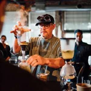 A male chef wearing safety goggles as he creates a steaming concoction. There are people behind him watching the process. Chef Todd Annis, Cru Catering