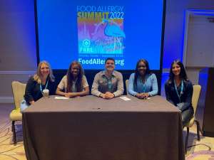 Five young professionals sitting behind a table with a brown covering and in front of a screen with the FARE Food Allergy Summit 2022 logo displayed