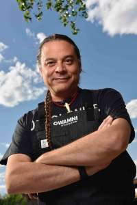 Smiling Native American man standing outside. He is wearing a black shirt and chef's apron, which has the Omamni by The Sioux Chef logo in top center printed. His arms are crossed and his braid of hair is over his right shoulder. Chef Sean Sherman