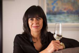 Woman with long dark hair wearing a black shirt leans on a high table while holding a glass of white wine Amanda Thomson, Thomson & Scott, Women Making HERStory promoting transparency in wine labeling
