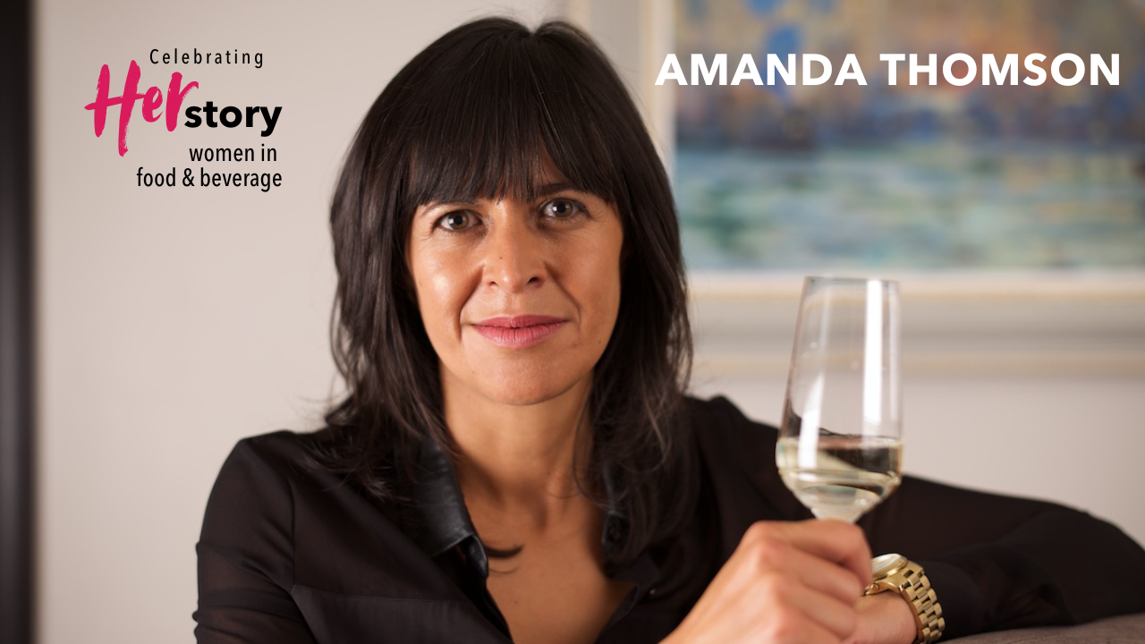 Woman with long dark hair wearing a black shirt leans on a high table while holding a glass of alcohol-free white wine Amanda Thomson, Thomson & Scott, Women Making HERStory with greater transparency in wine labeling.