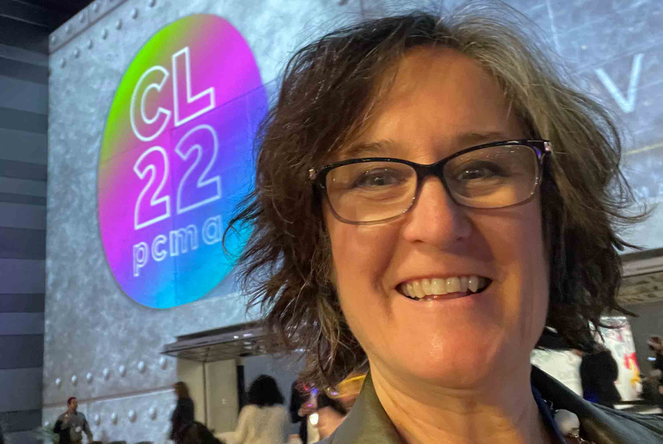 close-up selfie of smiling woman standing in front of a sign on a wall - CL22 PCMA