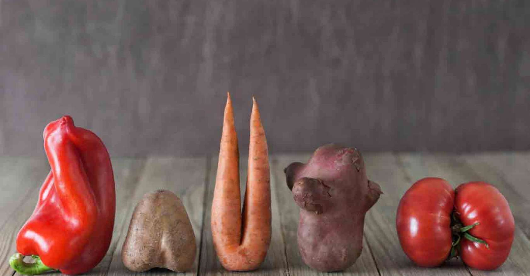 A bell pepper, potato, carrot with two "legs", sweet potato, and a tomato lined up on a wooden table with a brown wall behind them. They are representing ugly vegetables, imperfect vegetables.