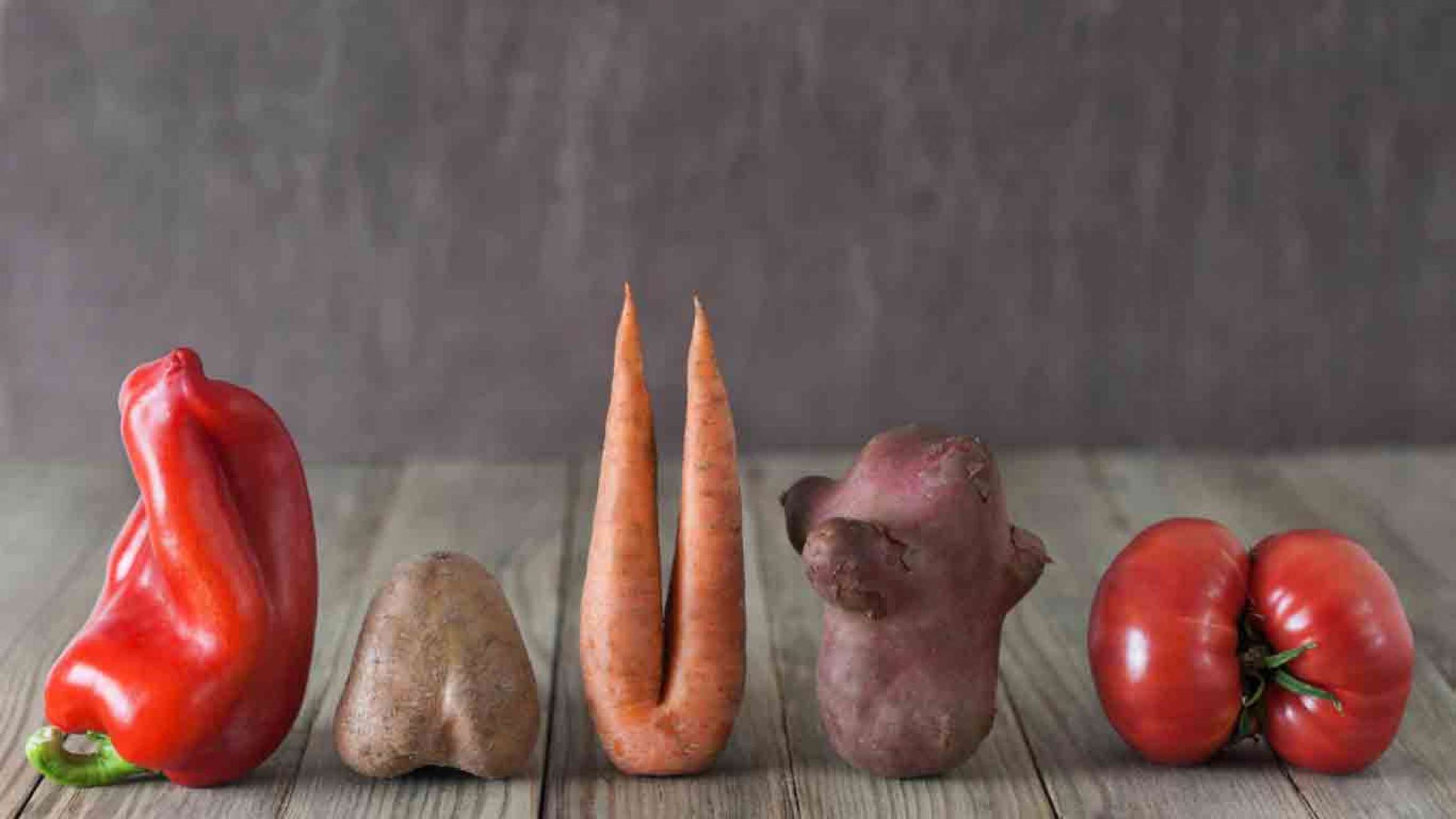 A bell pepper, potato, carrot with two "legs", sweet potato, and a tomato lined up on a wooden table with a brown wall behind them. They are representing ugly vegetables, imperfect vegetables.