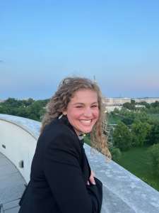 Young smiling woman in a black jacket leaning on a wall looking at the camera. The US Capital where she can be found Lobbying for Systemic Change in Food Service Elise Buellesbach