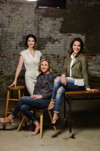 Three women co-founders of non-alcoholic beverage company Spiritless. One is standing wearing a cream colored dress. In front of her is a woman sitting n a chair with her left leg crossed over her right. The one in front is wearing jeans a white blouse and a jacket while sitting on a table with her left leg crossed over her right and her left hand on the table.