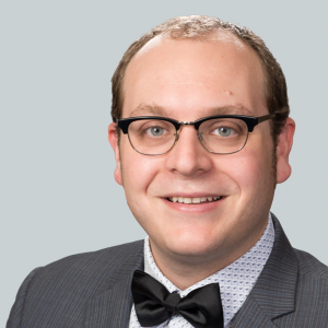 Smiling man with glasses looking at the camera. His dark hair is receding. He is wearing a gray suit with a patterned white shirt and a black bow-tie. His name is Andrew Hartley and he studies hotel food and beverage trends.
