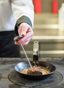 chef's hand holding a meat thermometer testing meat in a frying pan, ensuring it is following food safety practices