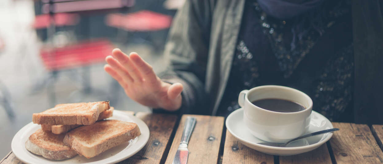 Person seated at a café table rejecting a plate of toasted bread, indicating a gluten-free diet; a cup of coffee sits in front of them on a rustic wooden table. This is in a post about Illinois celiac disease training bill SB 1288.