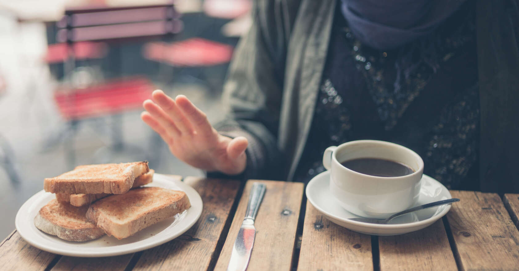 Person seated at a café table rejecting a plate of toasted bread, indicating a gluten-free diet; a cup of coffee sits in front of them on a rustic wooden table. This is in a post about Illinois celiac disease training bill SB 1288.