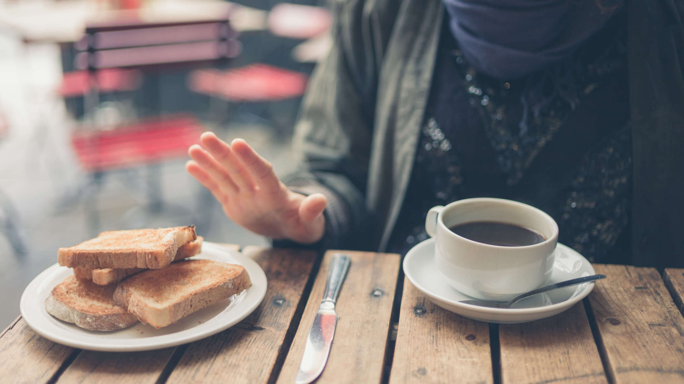 Person seated at a café table rejecting a plate of toasted bread, indicating a gluten-free diet; a cup of coffee sits in front of them on a rustic wooden table. This is in a post about Illinois celiac disease training bill SB 1288.