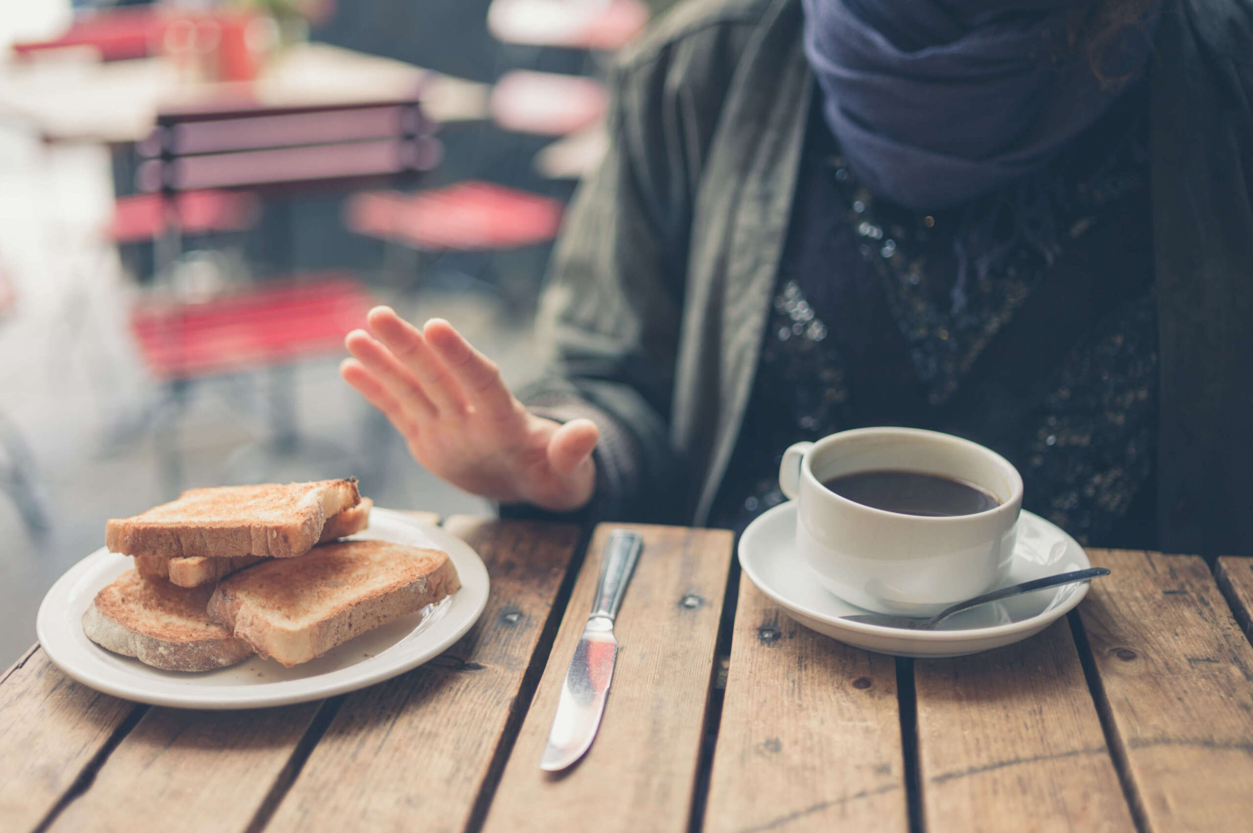 Person seated at a café table rejecting a plate of toasted bread, indicating a gluten-free diet; a cup of coffee sits in front of them on a rustic wooden table. This is in a post about Illinois celiac disease training bill SB 1288.