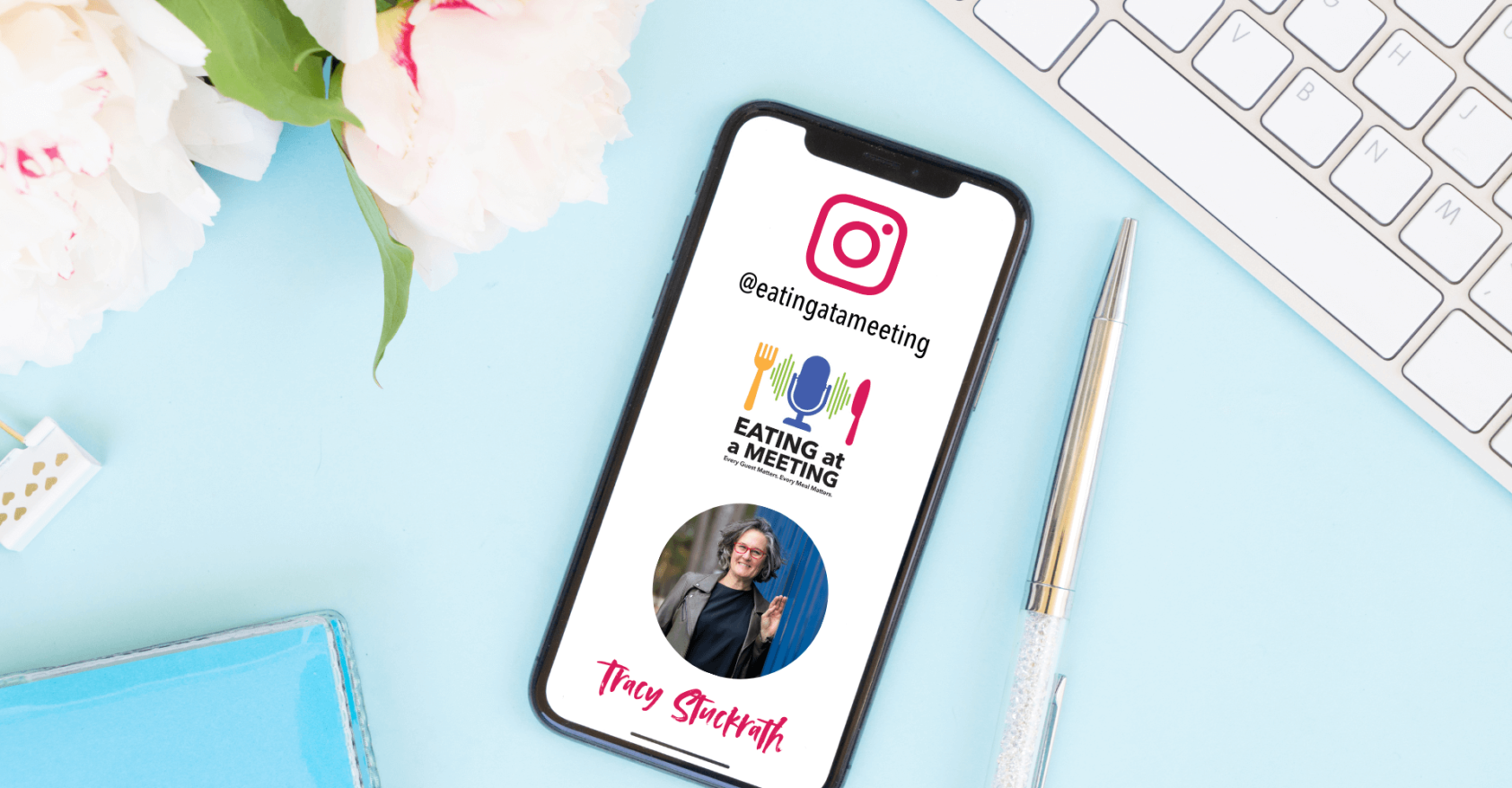A smartphone displaying the Instagram handle @eatingatameeting rests on a pastel blue desk beside a silver pen, white keyboard, and white peonies with pink edges. The phone screen features the Eating at a Meeting podcast logo, a circular photo of host Tracy Stuckrath, and her name in pink script at the bottom. The scene is bright and welcoming, styled with soft feminine touches.