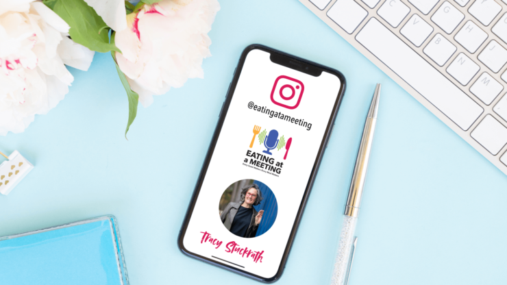 A smartphone displaying the Instagram handle @eatingatameeting rests on a pastel blue desk beside a silver pen, white keyboard, and white peonies with pink edges. The phone screen features the Eating at a Meeting podcast logo, a circular photo of host Tracy Stuckrath, and her name in pink script at the bottom. The scene is bright and welcoming, styled with soft feminine touches.