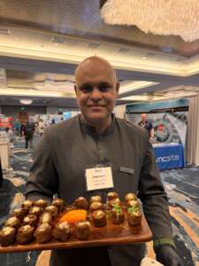 Server at a conference reception holding a wooden tray of crispy falafel bites topped with sauce and microgreens, labeled vegetarian and vegan, in a ballroom expo setting with booths in the background.