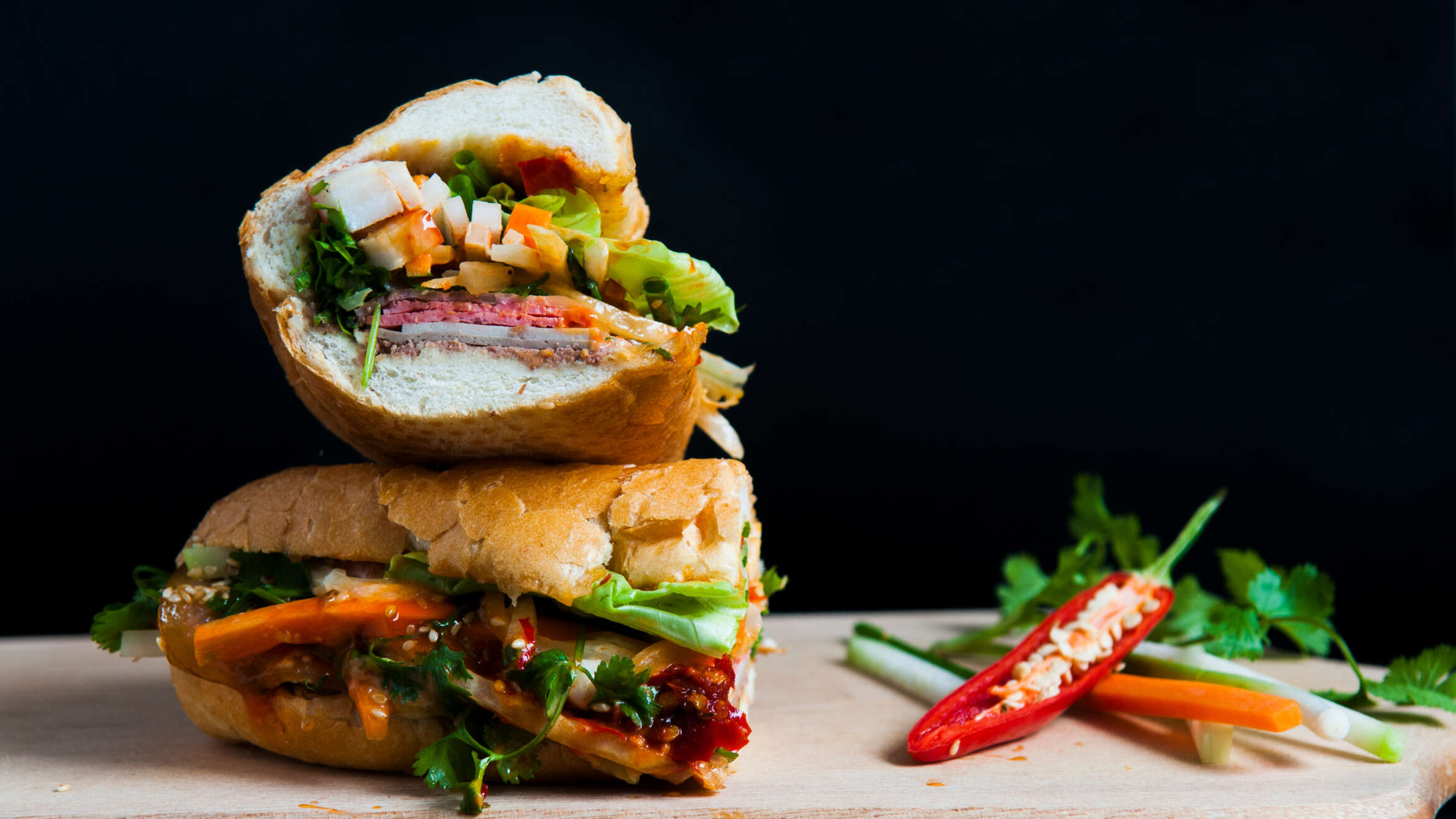 Two piece of a vegetarian Vietnamese sandwich stacked on top of each other on wooden cutting board. Two pepper sit to the right of the sandwich. This makes a great vegetarian dish option at meetings.