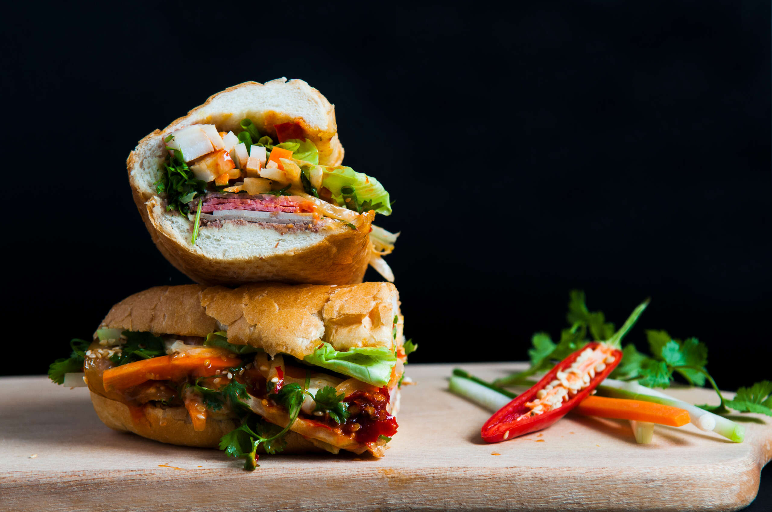 Two piece of a vegetarian Vietnamese sandwich stacked on top of each other on wooden cutting board. Two pepper sit to the right of the sandwich. This makes a great vegetarian dish option at meetings.