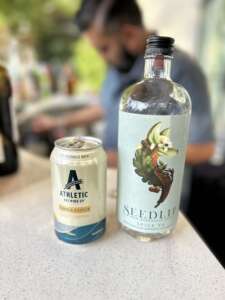 A can of Athletic NA beer sitting on an event bar next to a bottle of NA Seedlip. There is a bartender blurred out in the background.